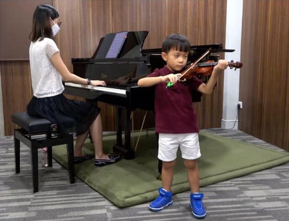 A woman playing the piano and a boy playing the violin. The boy is going through a violin examination.