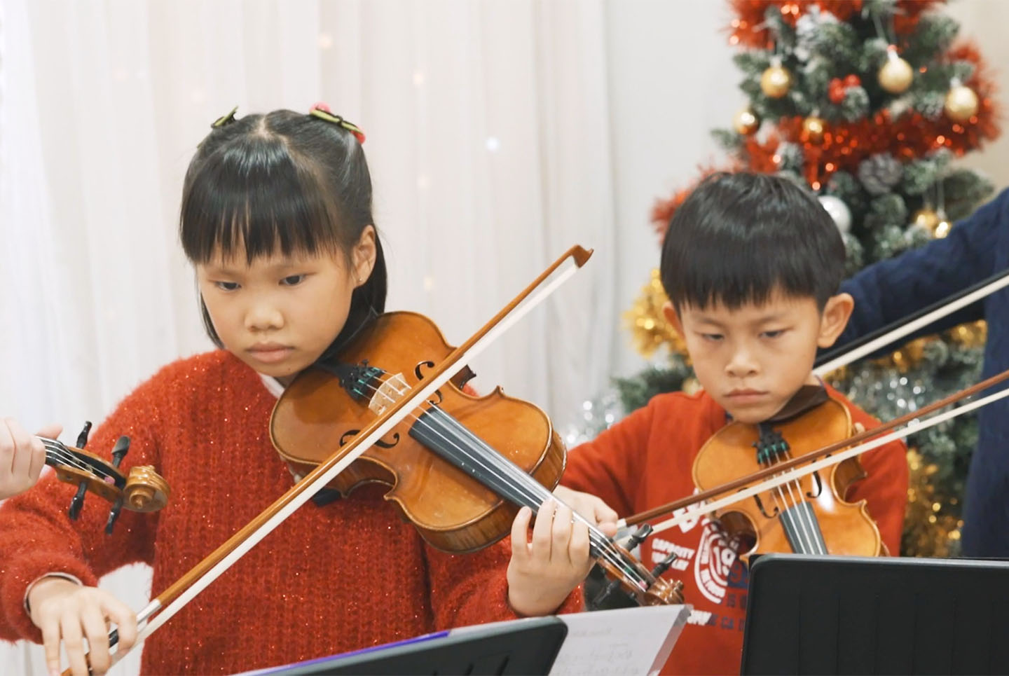 A photo of a boy and a girl dressed in red playing the violin next to a Christmas Tree